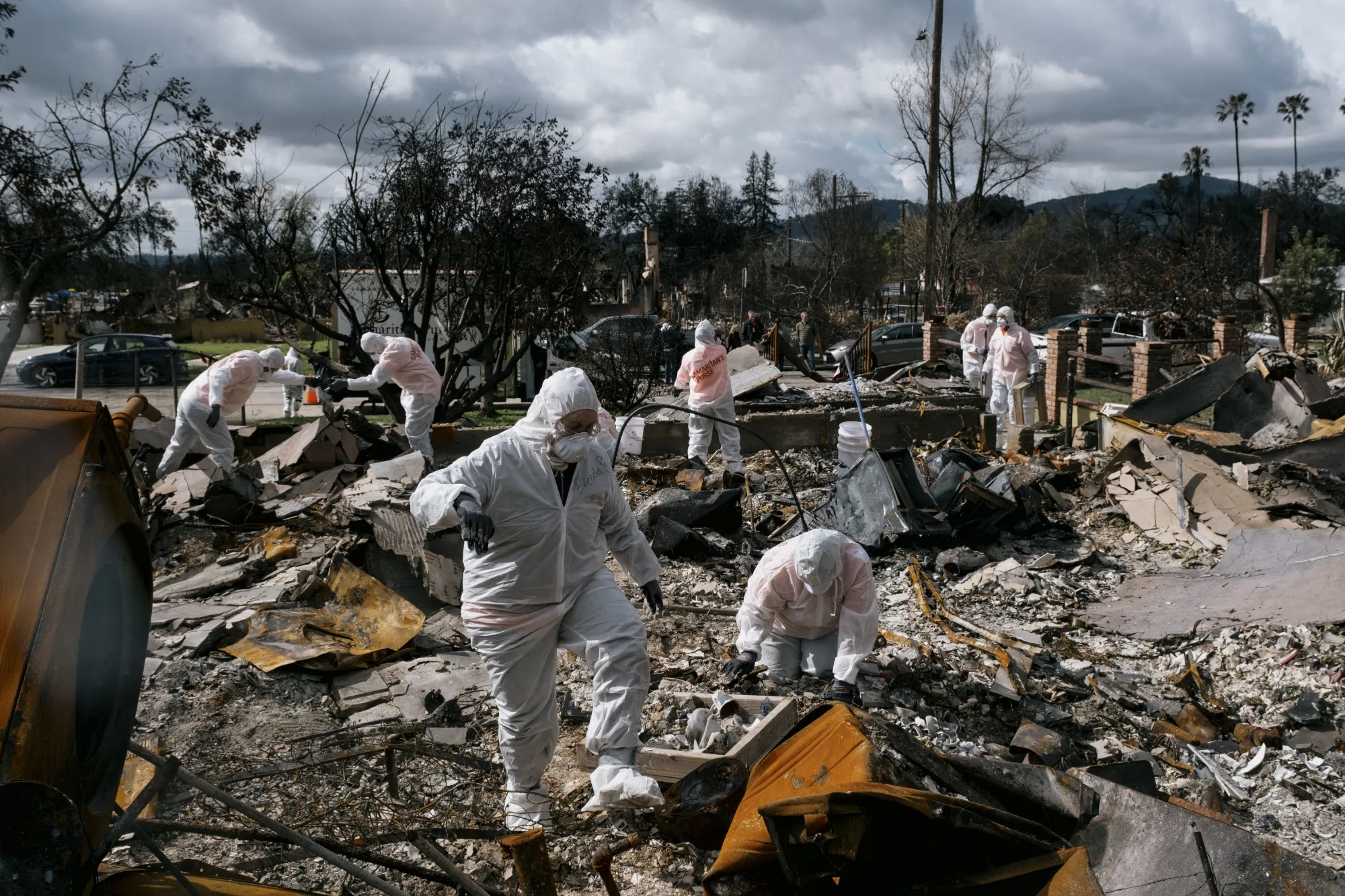 Volunteers in protective suits sifting through the debris of a burned neighborhood